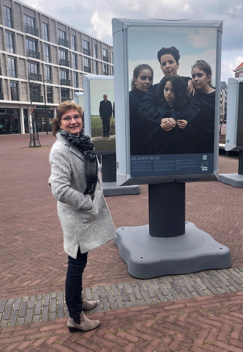 Ingrid van Sluijs bij de expositie. Ze staat bij een foto met daarop vier personen in donkere kleding. De middelste twee hebben een arm om elkaar heen geslagen.