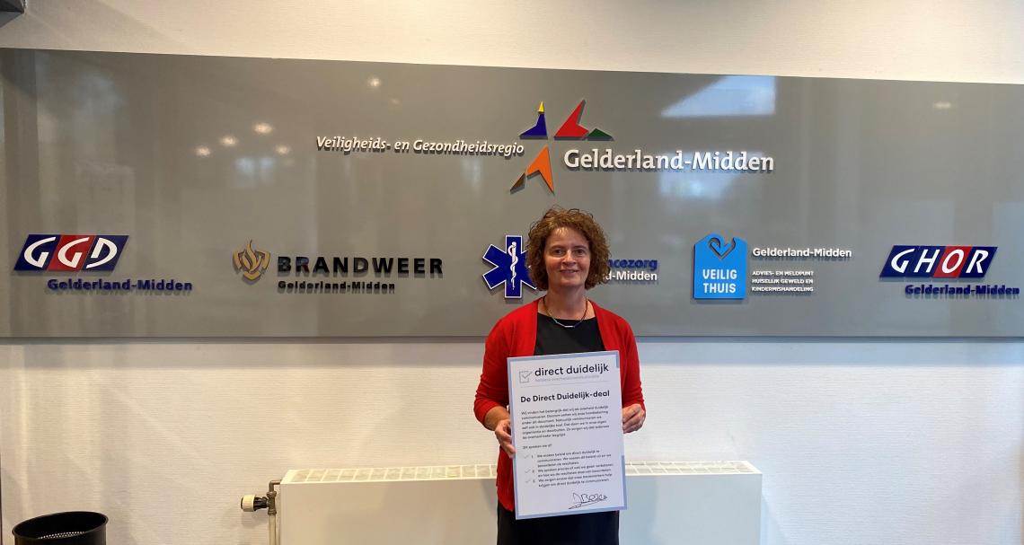 Janine Bezem, manager Jeugdgezondheidszorg (JGZ) van GGD Gelderland-Midden met het convenant in haar handen. Ze staat voor een muur met alle logo's van VGGM.